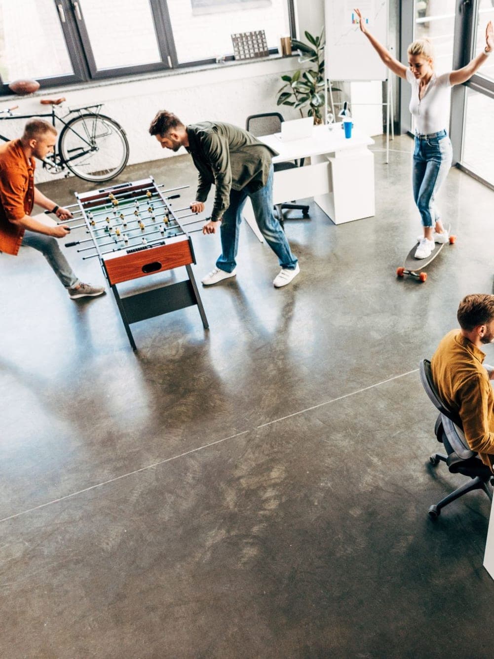 An open-plan office: colleagues at a foosball table, someone on a skateboard, two people talking over coffee